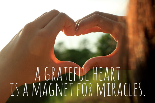 Inspirational quote  - A grateful heart is a magnet for miracles. With young woman hands making love sign against the sunset light. Thankfulness and gratefulness inspiration words concept.