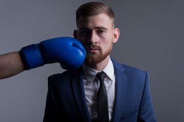 Naklejka premium close-up portrait of a young bearded guy in a business suit, businessman, side view the hand of an opponent in a boxing glove strikes. The moment of punching in the face.