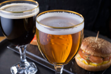 close-up, beer glasses with craft dark and light beer with foam, against a background of burgers, with a variety of ingredients, including shyomga, beef meat patty, cheese burger. In a rustic style