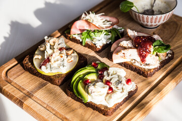 Four different rye bread open sandwiches (avocado-quail eggs, pear-gorgonzola, ham-cheese, turkey-apple) and bowl with mustard sauce on wooden board on white background.