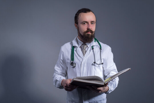 Portrait of a bearded doctor in a white coat and a phonendoscope holding educational books in his hands. Studio photo on a gray background.