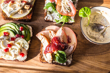 Four different rye bread open sandwiches (avocado-quail eggs, pear-gorgonzola, ham-cheese, turkey-apple) and bowl with mustard sauce on wooden board on white background.