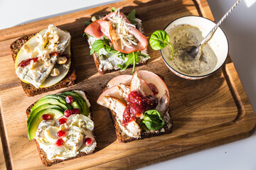 Four different rye bread open sandwiches (avocado-quail eggs, pear-gorgonzola, ham-cheese, turkey-apple) and bowl with mustard sauce on wooden board on white background.