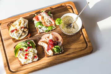 Four different rye bread open sandwiches (avocado-quail eggs, pear-gorgonzola, ham-cheese, turkey-apple) and bowl with mustard sauce on wooden board on white background.