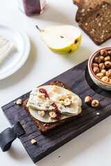 Open sandwich with rye bread, fresh pear , gorgonzola  cheese, hazelnuts and cranberry sauce and bowl with nuts on wooden board and sandwich ingredients  on white background. 