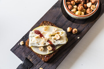 Open sandwich with rye bread, fresh pear , gorgonzola  cheese, hazelnuts and cranberry sauce and bowl with nuts on wooden board on white background. 