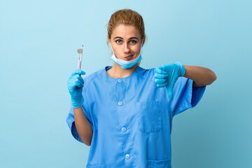 Young woman dentist holding tools over isolated blue background showing thumb down sign