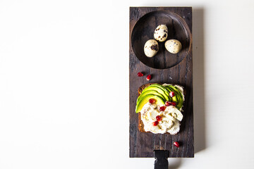 Open sandwich with rye bread, avocado, cream cheese, quail eggs and pomegranate seeds and three quail eggs  on wooden board on white background. 
