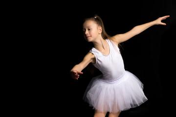 Naklejka premium Cute little girl in a tutu and pointe shoes is dancing in the studio on a black background.