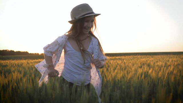 Pretty child in the hat is running across the wheat field. Happy young girl running in the field at sunset. Freedom concept.