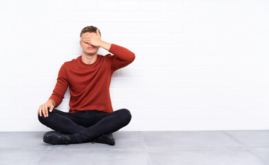 Young handsome man sitting on the floor covering eyes by hands