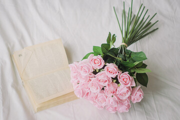 A bouquet of pale pink roses on a bed in the morning next to an open book. Reading a romantic novel.