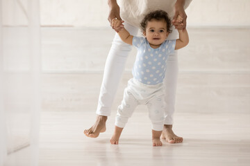 Caring barefoot African American mother teaching cute toddler daughter to walk, loving mum holding...