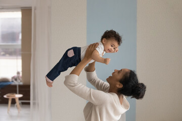 Laughing African American young mother playing with cute little daughter, holding funny baby pretending flying, loving happy mum lifting adorable toddler girl, having fun together at home