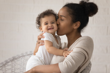 Close up loving young African American mother kissing cute toddler daughter cheek, sitting in cozy...