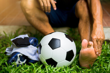 Soccer ball and sport shoes on green grass with soccer player in ankle pain. Soccer player sitting...