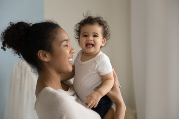 Smiling loving African American woman holding funny cute toddler girl, standing in nursery at home, happy young mum enjoying tender moment, leisure time with adorable little daughter