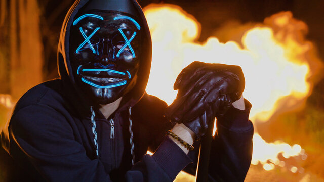 Criminal man in black hoodie and anonymous neon mask, leans on the baseball bat
