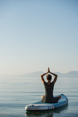 Woman meditating on sup board floating on calm sea water