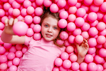 Fototapeta na wymiar A little happy girl in pink clothes with a smile lies in pink plastic balloons on a pink background. The view from the top.