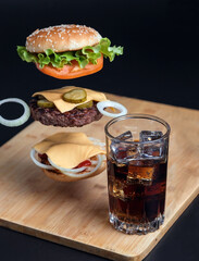 Delicious Burger with floating ingredients on the background of a wooden table. Bun with sesame seeds. Juicy meat cutlet. Carbonated brown drink with ice.