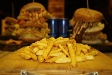 A plate of french fries. In the background there is a group of American sandwiches, consisting of burger slices, cheese, and fried chicken.