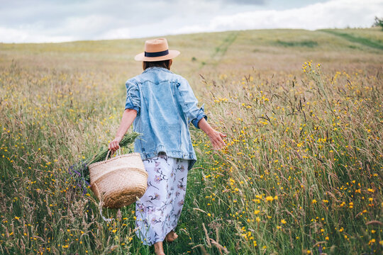 Young Woman dressed jeans jacket and light summer dress walking by the high green grass meadow with basket and wildflowers bouquet. Human and nature concept back shot photo.