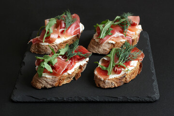 Buckwheat toasts with goat cheese, prosciutto and arugula lying on a black stone board on a black table.