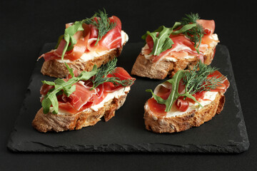 Buckwheat toasts with goat cheese, prosciutto and arugula lying on a black stone board on a black table.