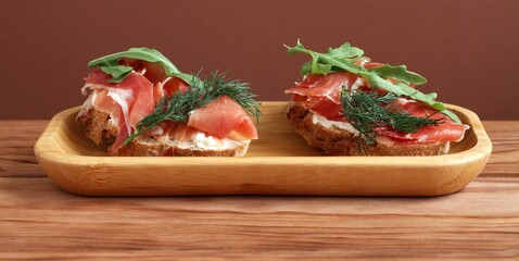 Buckwheat toasts with goat cheese, ham and arugula lying on a bamboo plate on a wooden table.