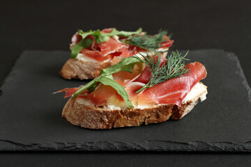 Buckwheat toasts with goat cheese, prosciutto and arugula lying on a black stone board on a black table.