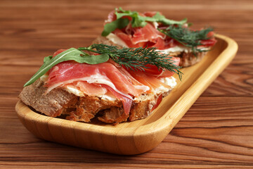 Buckwheat toasts with goat cheese, ham and arugula lying on a bamboo plate on a wooden table.