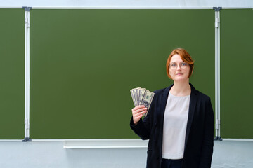 School teacher holds money in us dollars and smiles on blackboard background, copy, space