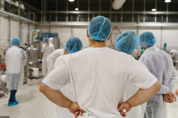 Back view on group of people wearing protective bouffant caps and masks standing and watching the production process at factory - High demanding health hygiene and standard safety concept - obrazy, fototapety, plakaty