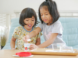 Little Asian baby girl ( right ) smiling and enjoys scooping her little sister's ice cream - siblings rivalry / taking advantage