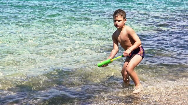 Slow motion of caucasian toddler boy, playing with a water gun, at beach of Sesi, Attica, Greece.