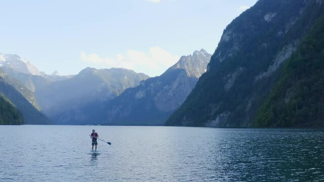 Aerial view Panning shot, Man in red shirt and cap paddle boarding in the middle of the lake surrounded by mountains in the background.