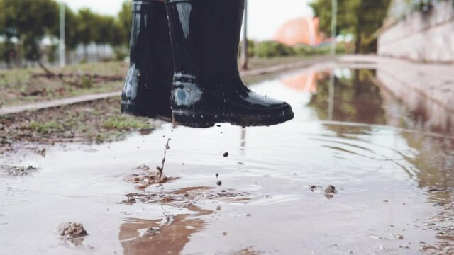 Close up video of kid  feet wearing rubber boots jumping in a muddy puddle on a rainy day at the park. Boy splashing and playing with water outdoors. Typical childhood fun memories