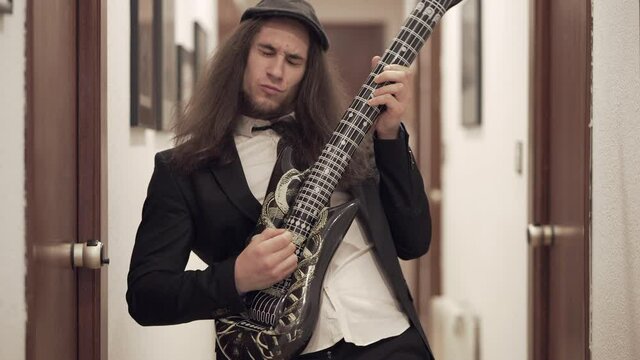 Young long-haired man in suit plays inflatable toy guitar in apartment