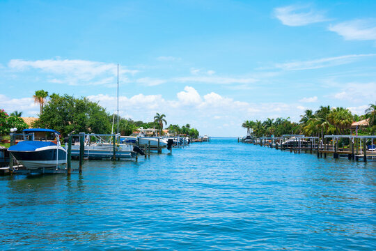 Residential saltwater canal in south Florida with beautiful blue water and a variety of boats at docks and on electric lifts with green palm trees lining the shoreline on a bright sunny afternoon.
