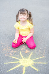 Smiling girl with syndrome down draws the sun with chalk on the asphalt