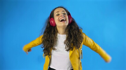 Young woman in yellow jacket dancing while listening music on headphones in Studio with blue...