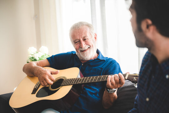 happy adult son and senior father playing guitar on sofa at home relaxing and having happy time with family