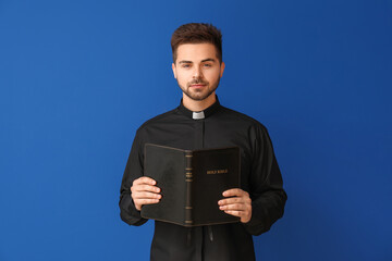 Young priest with Bible on color background