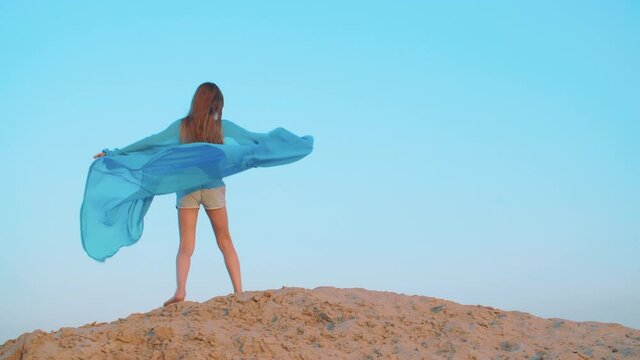 Playful girl teenager in waving blue pareo walking on sandy hill in wind summer desert. Cheerful girl in blue dress twisting on peak sandy dune in hot desert on sky landscape