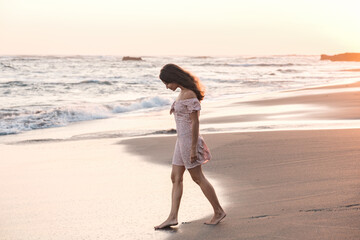 Young beautiful woman in a dress goes to the ocean along the beach at sunset. Girl with her head...