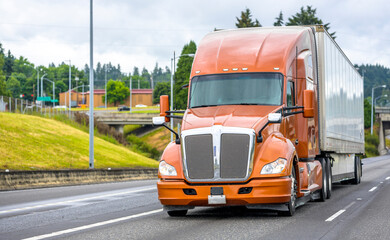 Fototapeta premium Bright brown big rig bonnet long haul semi truck transporting cargo in dry van semi trailer moving on the straight wide highway road with bridge and hill