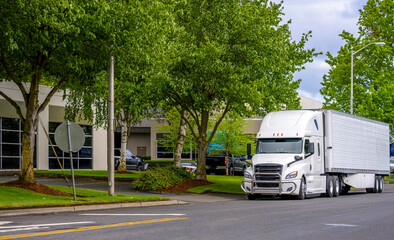 Stylish big rig white bonnet semi truck with refrigerator semi trailer for transportation frozen cargo standing on the city road with green trees and warehouse buildings