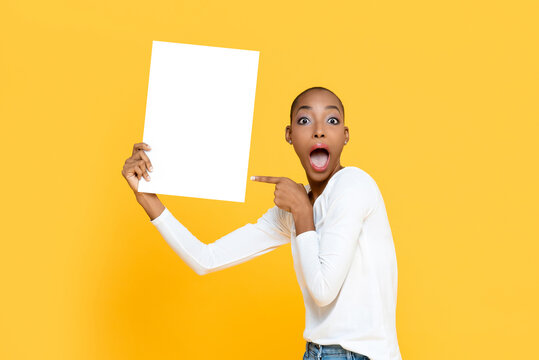 Portrait of surprised young African American woman with mouth open holding and pointing to white placard in isolated studio yellow background