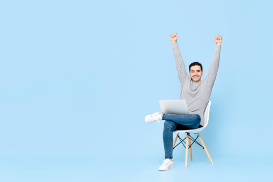 Portrait of happy young handsome Caucasian man sitting using laptop doing arms raised winning gesture in isolated studio blue background